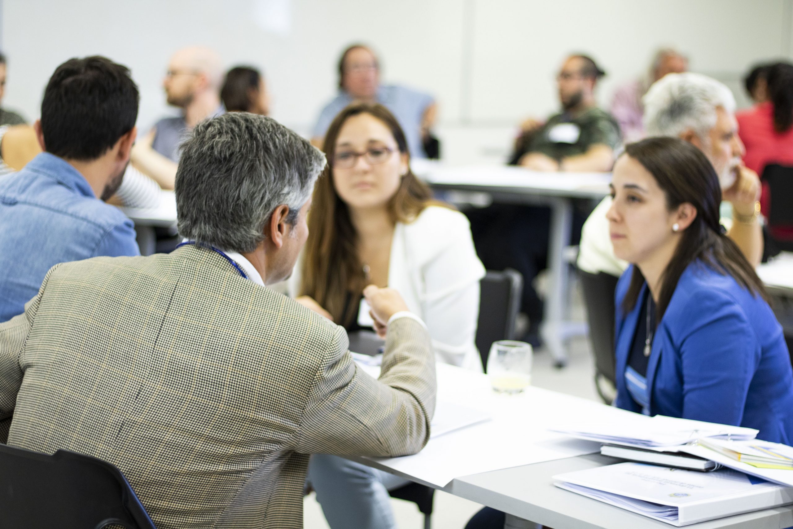 Docentes sentados conversando en una sala con otros docentes de fondo